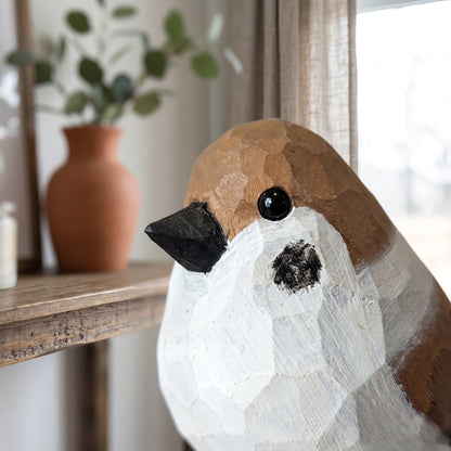 A close-up of a hand-painted wooden Sparrow sculpture, likely a robin, with a brown and white textured finish, black beak, and black eye, set on a wooden shelf next to a terracotta vase with eucalyptus.
