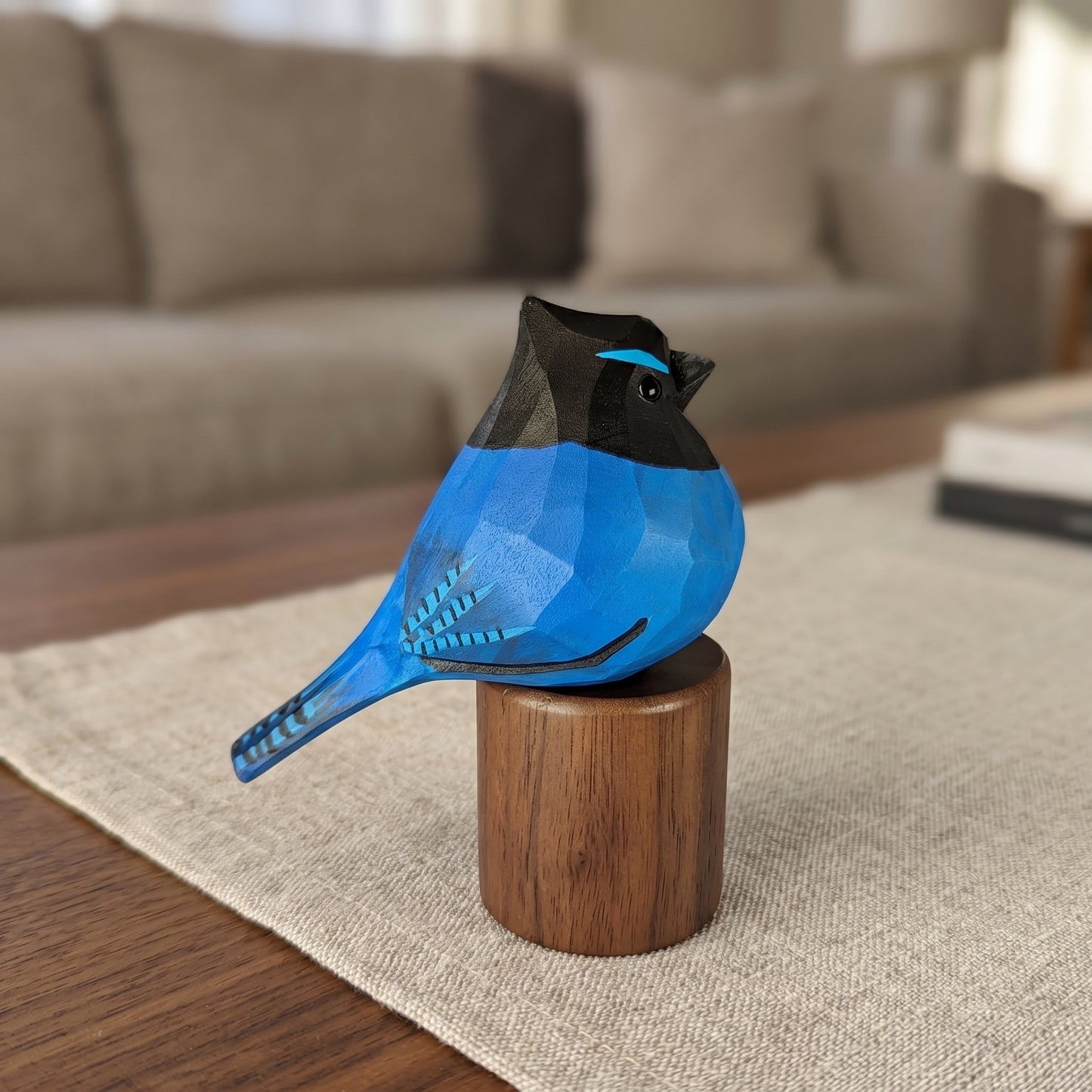 A carved wooden stellers jay figurine in bright blue and black, mounted on a small wooden pedestal, set on a table runner in a softly blurred living room.