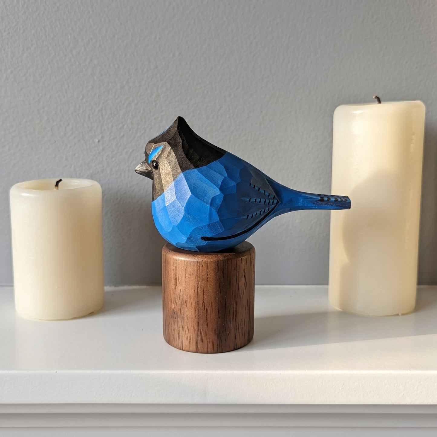 A carved wooden stellers jay sculpture in shades of blue, black, and gray, perched on a cylindrical wooden base. The sculpture is displayed on a white shelf between two tall cream-colored pillar candles against a gray wall.