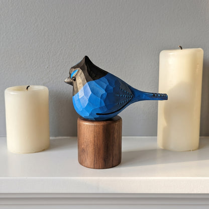 A carved wooden stellers jay sculpture in shades of blue, black, and gray, perched on a cylindrical wooden base. The sculpture is displayed on a white shelf between two tall cream-colored pillar candles against a gray wall.