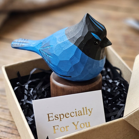 A small geometric stellers jay figurine in blue, black, and gray sits on a wooden base inside an open kraft gift box with black shredded paper and a card that reads 'Especially For You.' The box is placed on a rustic wooden surface with blurred natural background elements.
