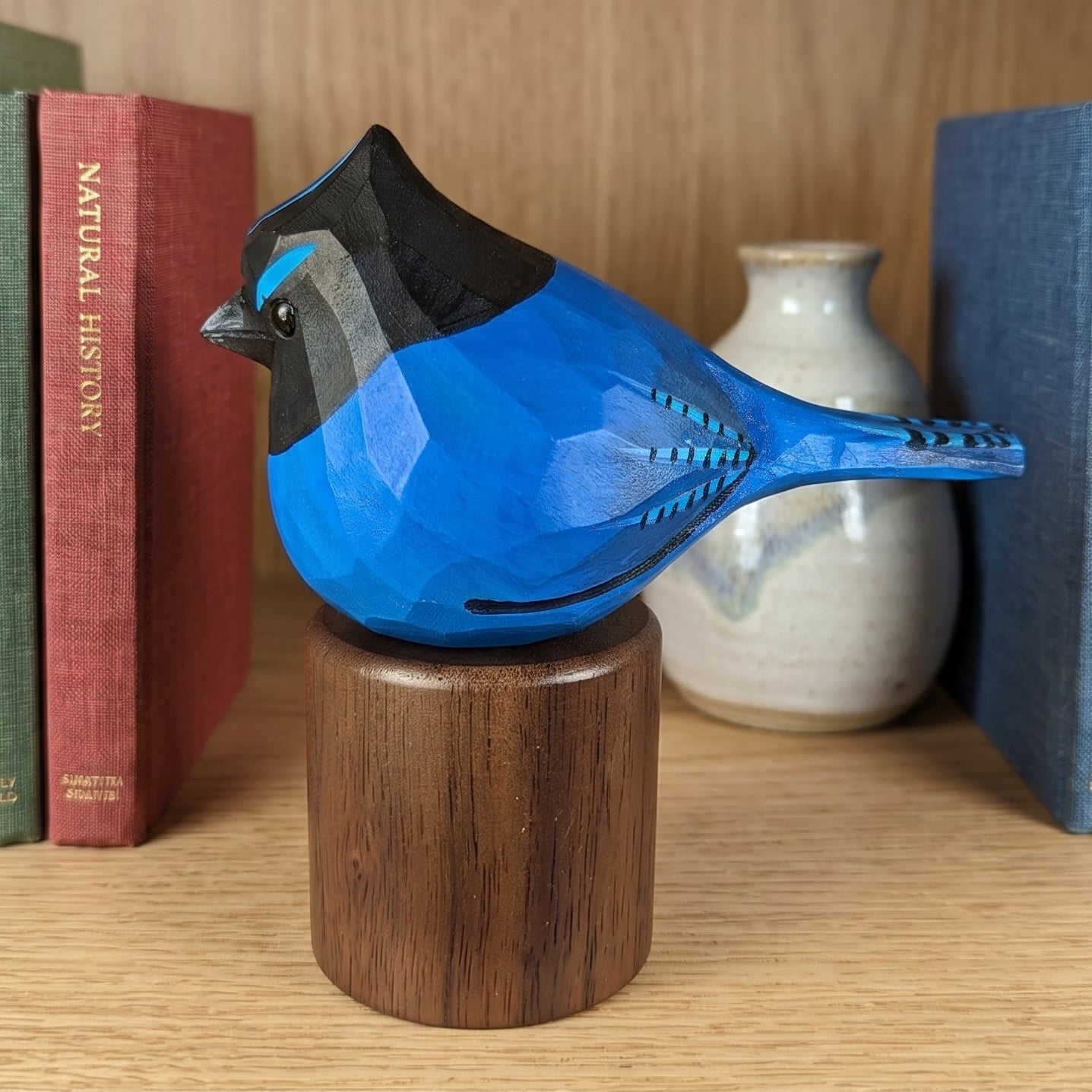 A geometric, faceted blue stellers jay figurine with black and gray detailing, mounted on a round wooden base and displayed on a wooden bookshelf between books.