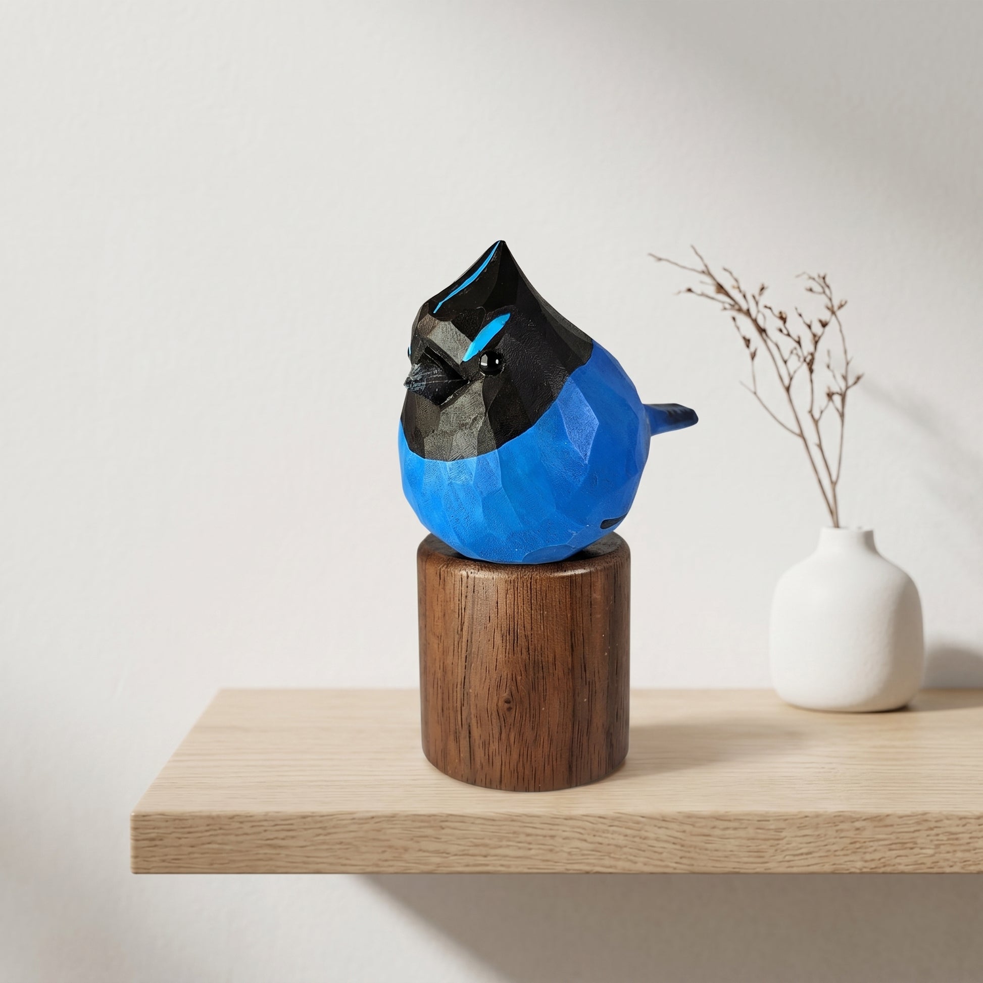 A faceted blue and stellers jay sculpture displayed on a dark wooden cylindrical pedestal, set on a light wood shelf next to a small white vase with dried branches.