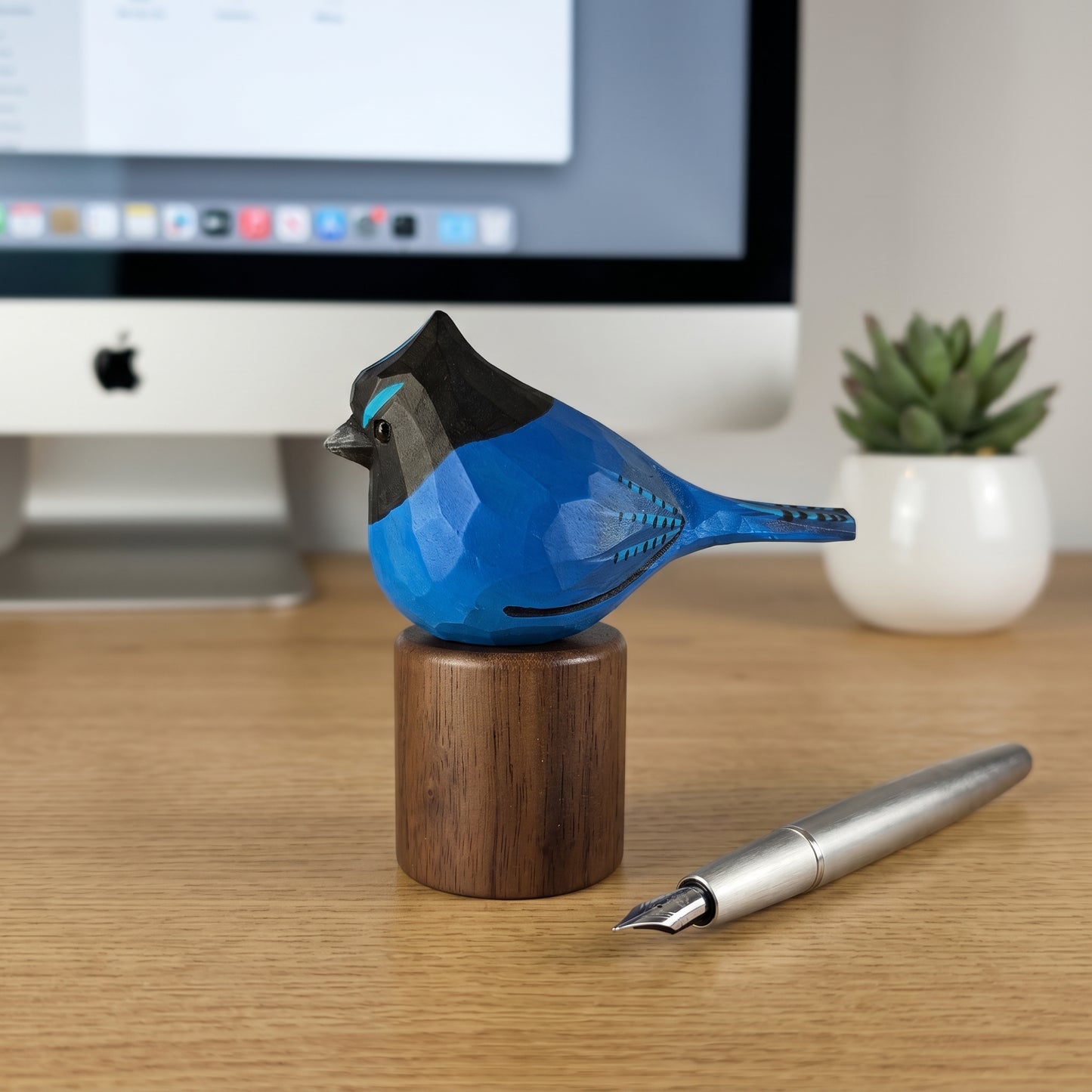 A hand-carved wooden stellers jay figurine in blue, black, and brown, displayed on a matching wooden stand on a desk next to a silver fountain pen, with an iMac and a small potted succulent in the background.