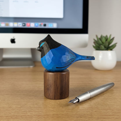 A hand-carved wooden stellers jay figurine in blue, black, and brown, displayed on a matching wooden stand on a desk next to a silver fountain pen, with an iMac and a small potted succulent in the background.