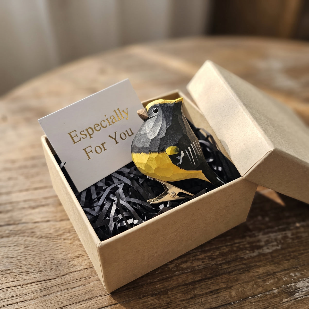 A small geometric Sultan Tit figurine in yellow, black, and gray sits in a kraft gift box with black shredded paper and a card that reads 'Especially For You,' placed on a wooden table.