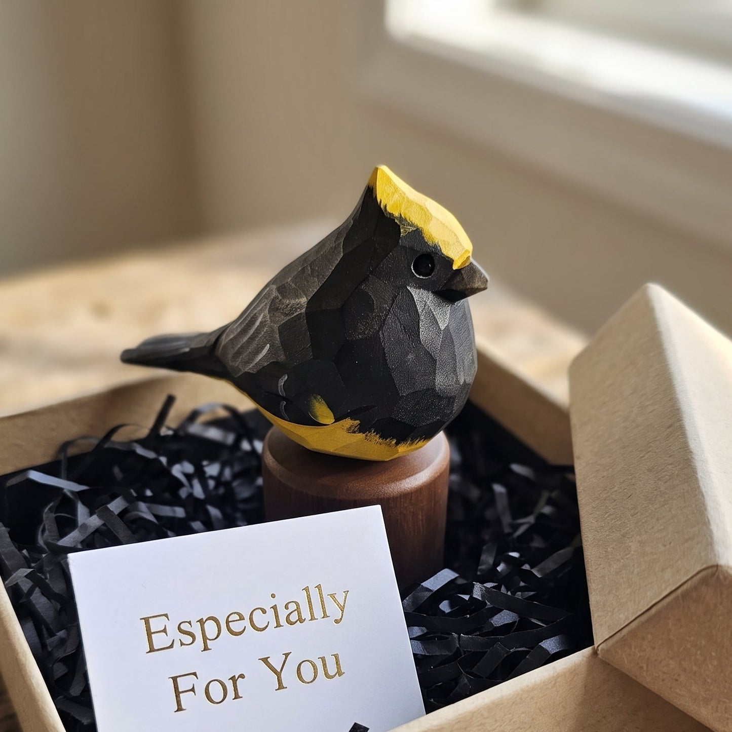A small geometric Sultan tit figurine with a yellow crest and black body sits in a kraft gift box filled with black shredded paper and a white card that reads 'Especially For You.' The box is placed on a rustic wooden surface near a window.