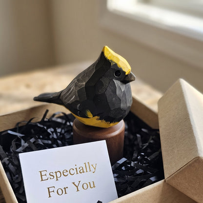 A small geometric Sultan tit figurine with a yellow crest and black body sits in a kraft gift box filled with black shredded paper and a white card that reads 'Especially For You.' The box is placed on a rustic wooden surface near a window.