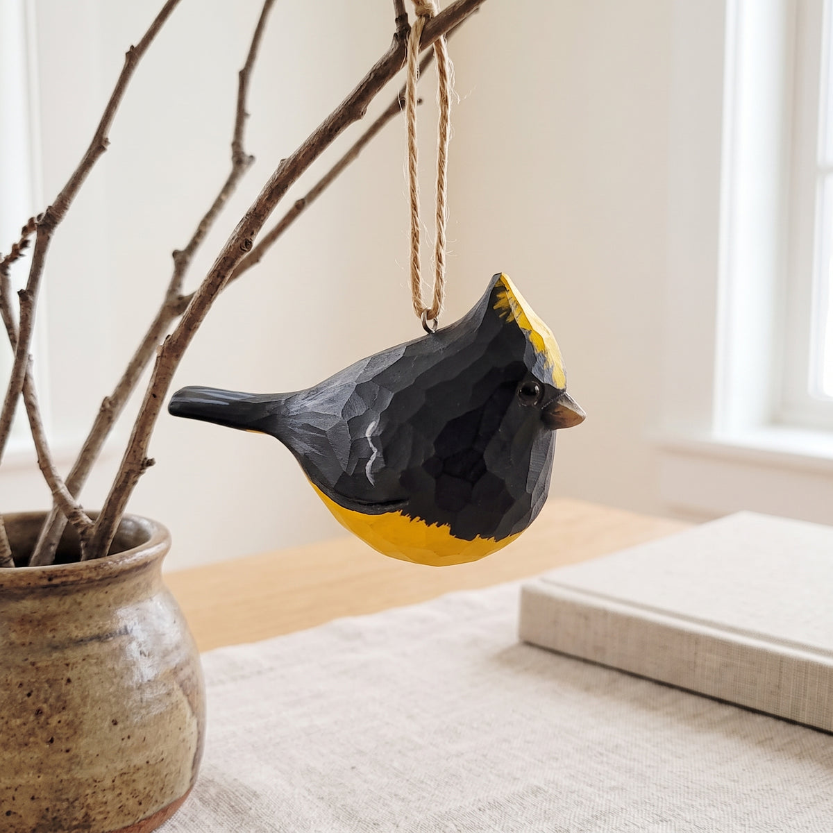 Hand-carved wooden Sultan Tit bird ornament hanging from branches in a ceramic vase near a brightly lit window