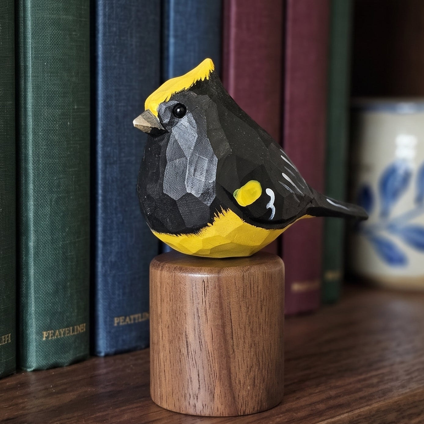 A hand-carved wooden Sultan tit figurine with a yellow and black patterned body, mounted on a round wooden base and displayed on a wooden bookshelf next to books and a ceramic mug.