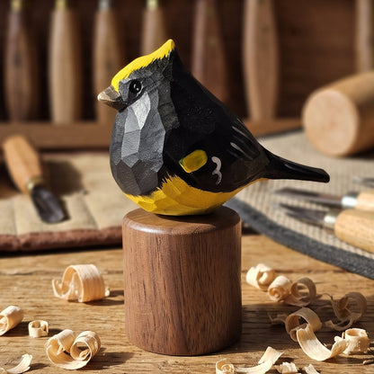 A handcrafted geometric Sultan tit sculpture in black and yellow wood, mounted on a cylindrical wooden base on a workbench with wood shavings, carving tools, and a mallet in the background.