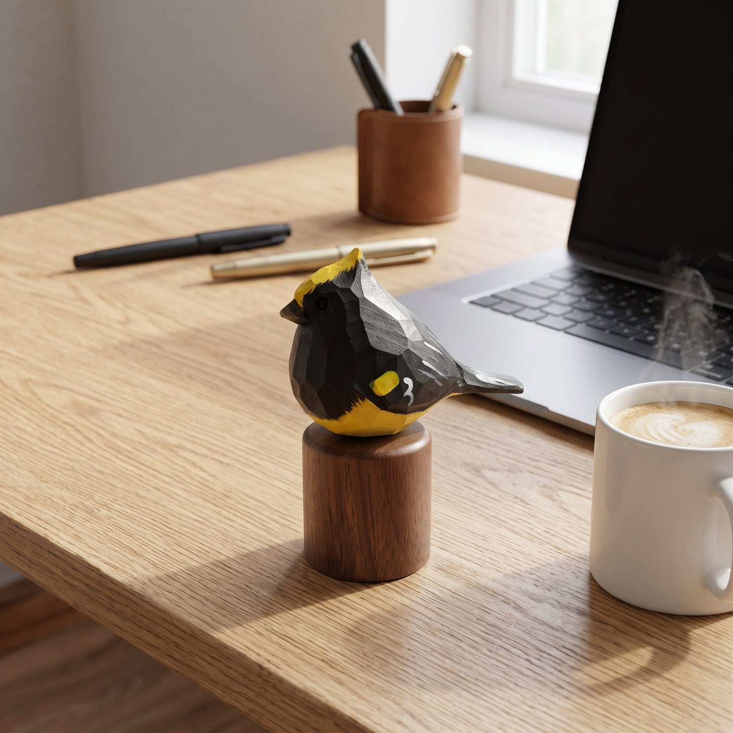 A small Sultan tit figurine with a black and yellow geometric pattern sits on a round wooden base on a light wood desk, with a laptop, coffee mug, and pen cup in the background.