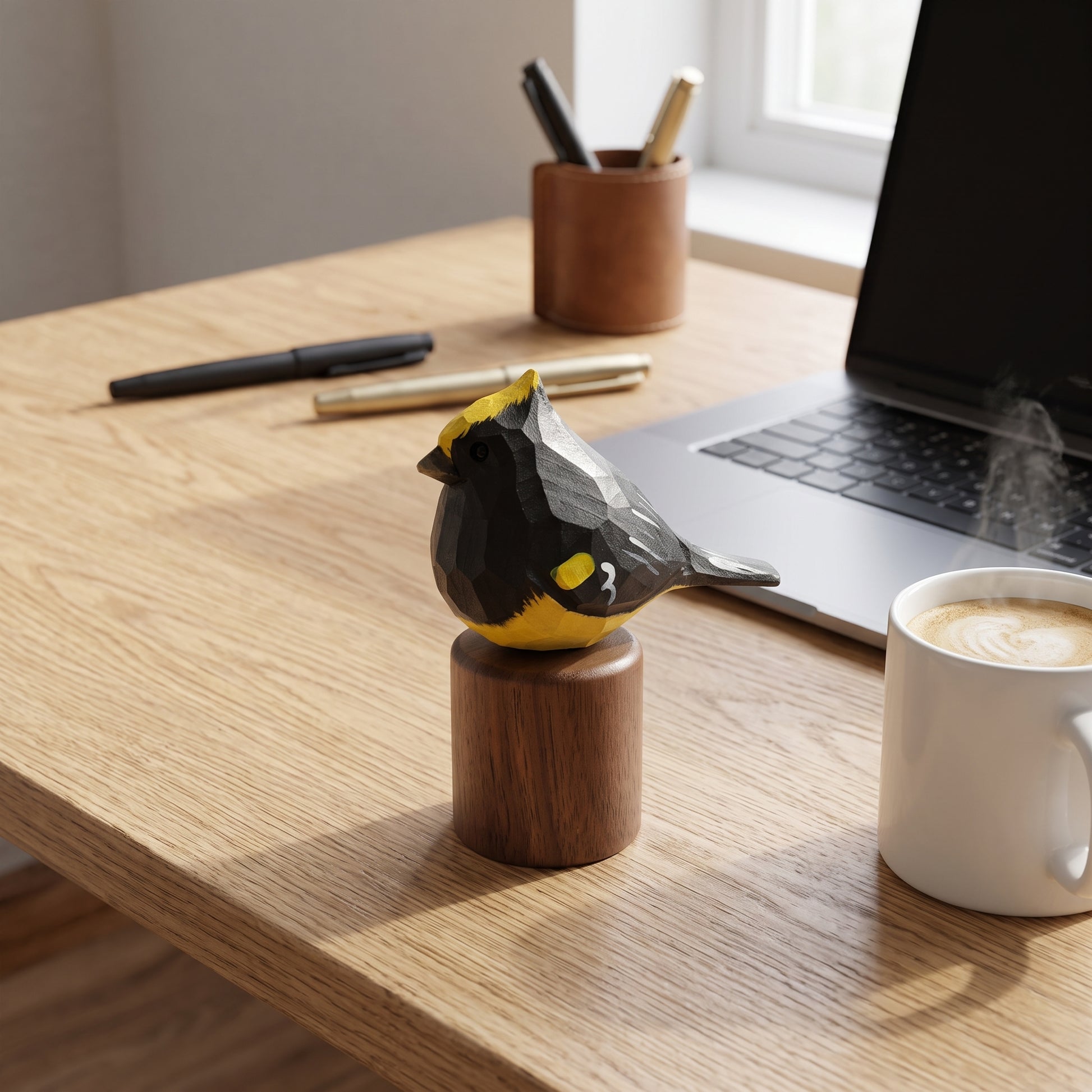 A small Sultan tit figurine with a black and yellow geometric pattern sits on a round wooden base on a light wood desk, with a laptop, coffee mug, and pen cup in the background.