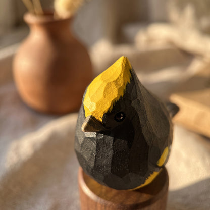 A hand-carved wooden Sultan tit figurine with a black and yellow painted finish, displayed on a wooden stand in a warm, softly lit setting.