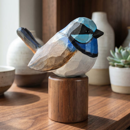 Hand-carved wooden Superb Fairywren figurine with bright blue plumage and black facial markings, mounted on a Black Walnut base on a sunlit wooden shelf.
