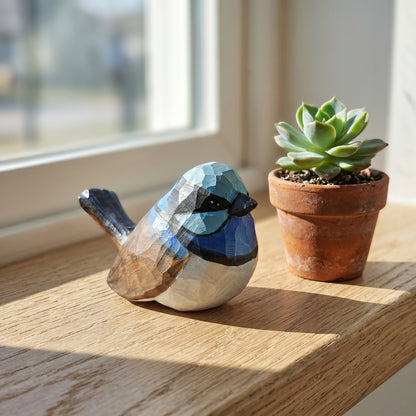 A geometric Superb fairywren figurine with blue, black, brown, and white facets sits on a wooden windowsill next to a small potted succulent.
