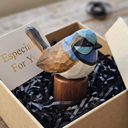 A handcrafted wooden Superb fairywren figurine with geometric facets sits in a brown kraft gift box filled with black paper shred and a card that reads 'Especially For You'. The box is placed on a rustic wooden tabletop.