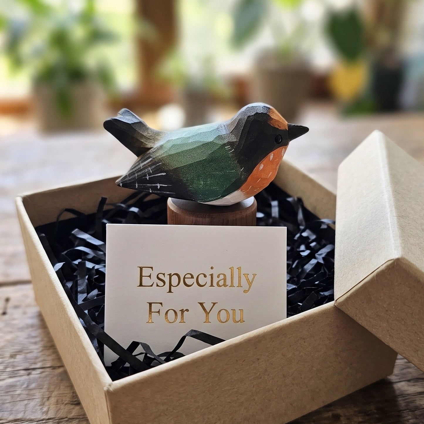 A small hand-painted Swallow figurine with green, orange, and black feathers sits in a brown kraft gift box filled with black paper shred and a card that reads 'Especially For You'. The box is placed on a rustic wooden table with a softly blurred indoor background featuring plants.