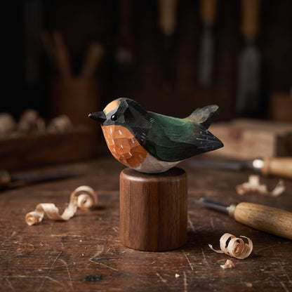 A handcrafted wooden Swallow figurine with a geometric patterned chest and a dark green back, displayed on a cylindrical wooden base on a rustic workbench with carving tools and wood shavings.