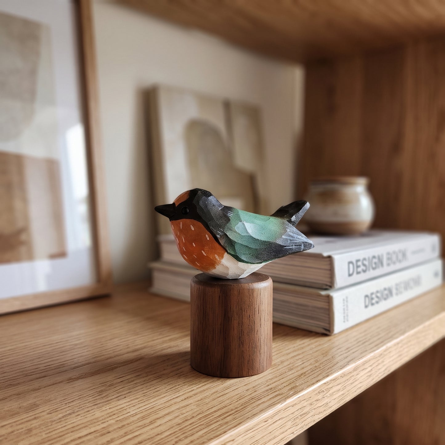 A small handcrafted wooden Swallow figurine with a black head, orange chest, and green wings, perched on a cylindrical wooden base on a light wood shelf.