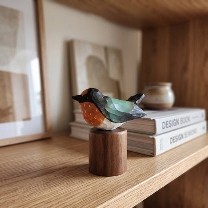 A small handcrafted wooden Swallow figurine with a black head, orange chest, and green wings, perched on a cylindrical wooden base on a light wood shelf.