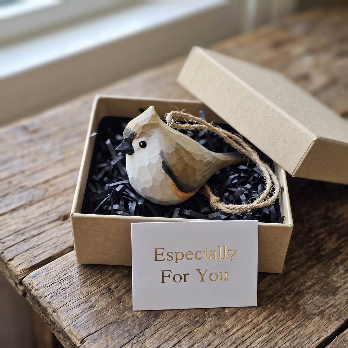 A faceted Tufted titmouse ornament in beige, brown, and black sits inside a kraft gift box with black paper shred and a card reading 'Especially For You' on a rustic wooden table.