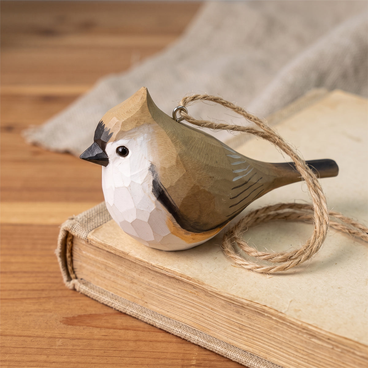 A handcrafted wooden Tufted titmouse ornament with a geometric faceted finish, resting on an old book with a loop of twine for hanging. The bird features a white face, a brown and beige back, and a black beak, set against a rustic wooden background.