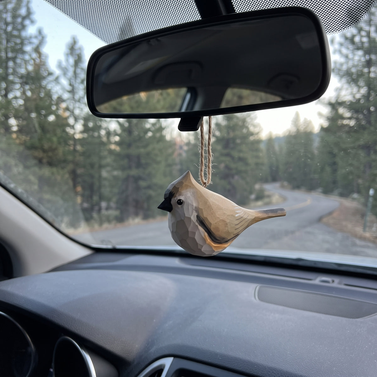 A small Tufted titmouse car air freshener hanging from a rearview mirror inside a vehicle, with a forest road visible through the windshield.