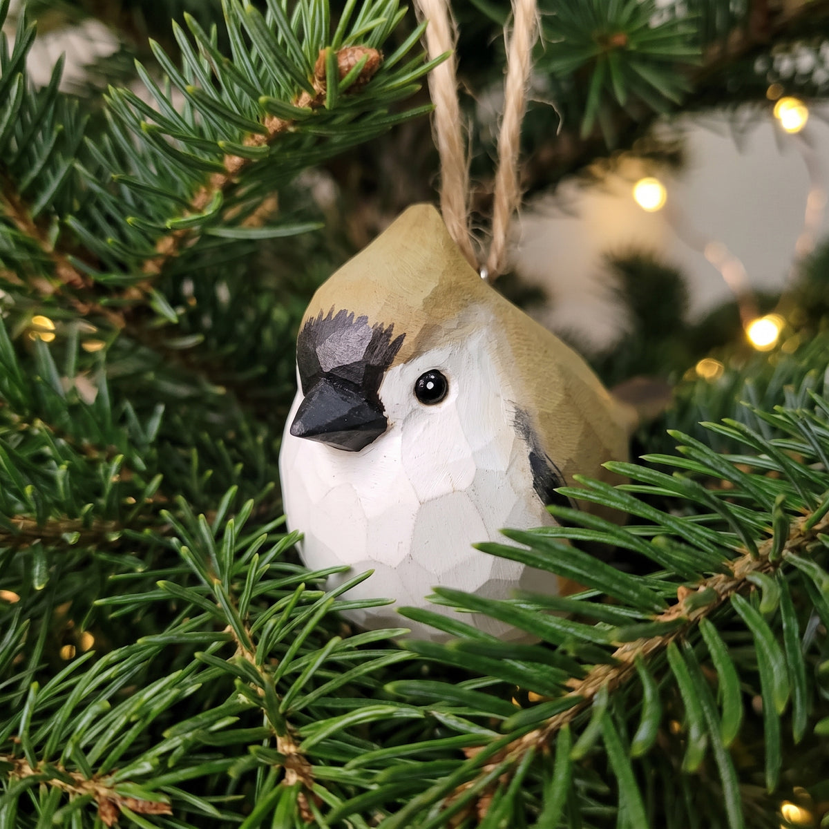 A handcrafted Tufted titmouse ornament shaped like a small bird with a tan head, white body, and black facial markings, hanging among green pine branches with warm string lights in the background.