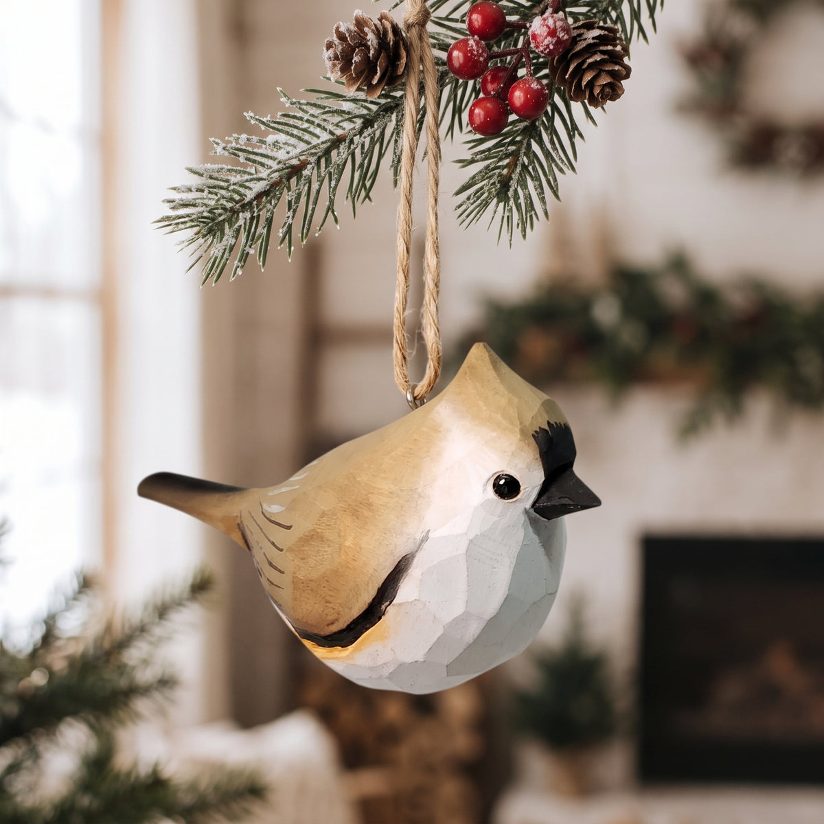 A geometric Tufted titmouse ornament shaped like a small cardinal or robin hangs from a pine branch with a twine loop. The ornament has a faceted, low-poly design with a tan back, white belly, and black beak, set against a softly blurred indoor background with holiday decor.