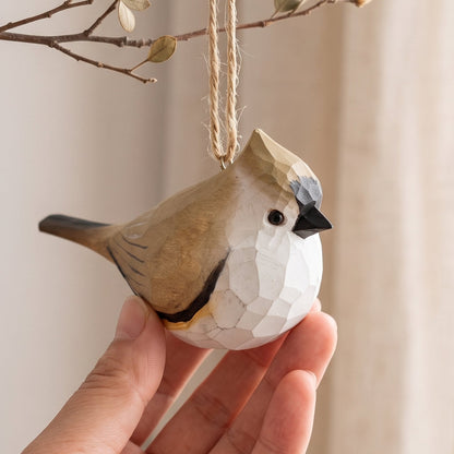 A hand holds a small Tufted titmouse -shaped ornament with a faceted geometric design. The ornament is tan and white with a black beak and a gray crest, hanging from a twine loop attached to a small branch.