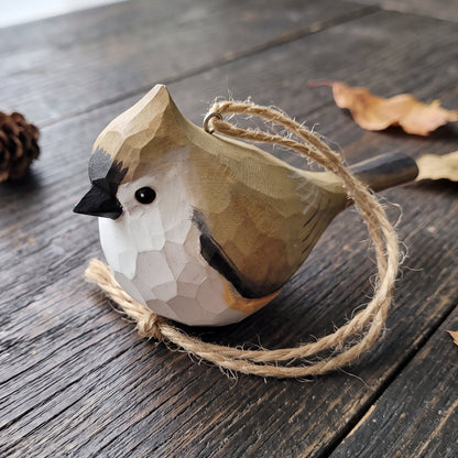 A carved wooden Tufted titmouse ornament with a natural wood hanging twine loop, resting on a dark rustic wooden tabletop with scattered dry leaves and a pinecone.