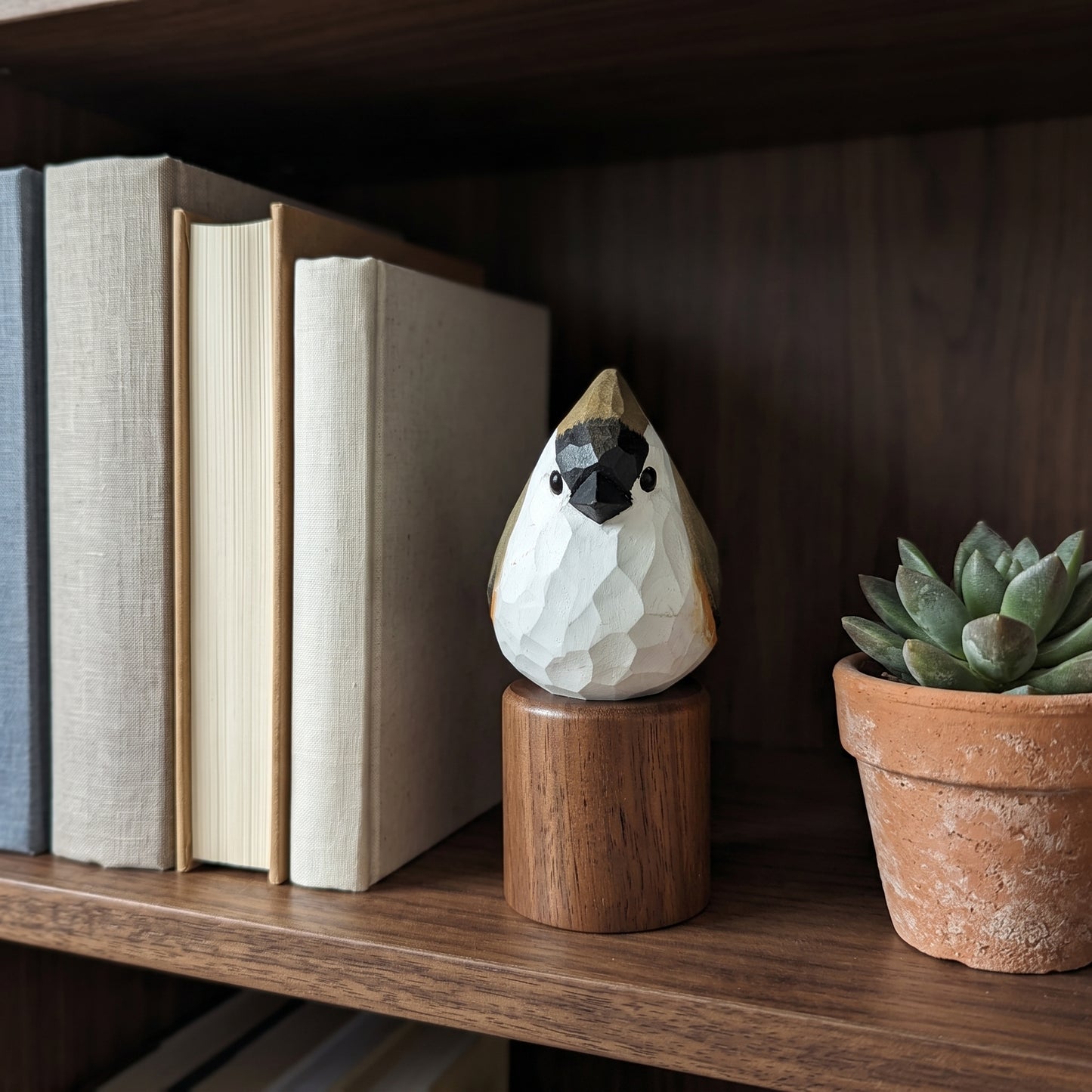 A small faceted Tufted Titmouse figurine in white, black, and gold sits on a wooden cylinder base on a dark wood bookshelf, with books and a potted succulent nearby.