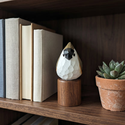 A small faceted Tufted Titmouse figurine in white, black, and gold sits on a wooden cylinder base on a dark wood bookshelf, with books and a potted succulent nearby.