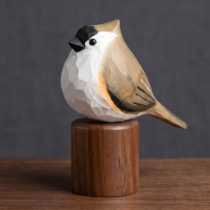 A hand-carved wooden Tufted Titmouse sculpture with a faceted geometric design, perched on a cylindrical wooden base. The bird features a white chest, black beak and back, and warm brown and beige tones on its wings and back.