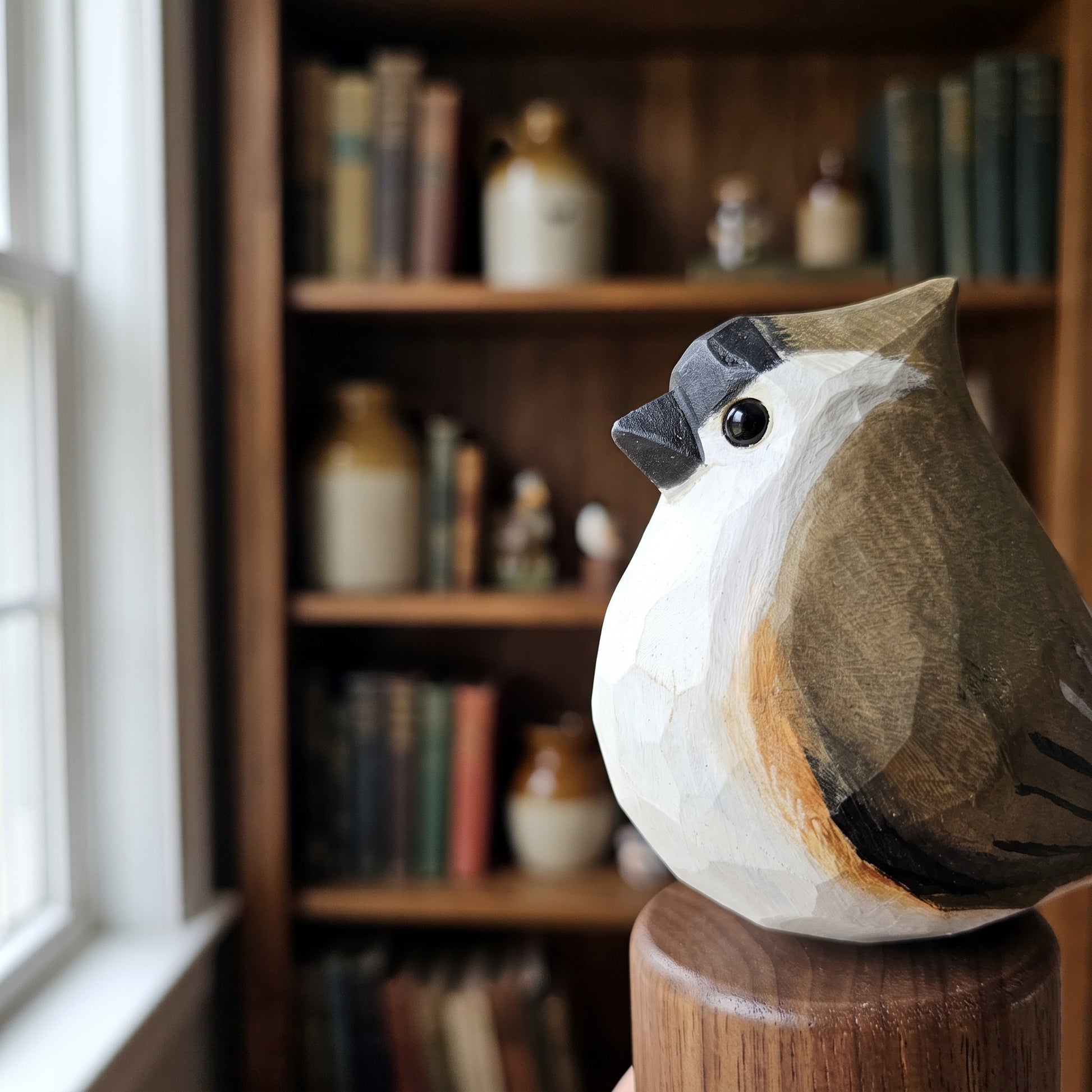 A hand-carved wooden Tufted Titmouse figurine with a white, brown, and black painted finish, perched on a wooden pedestal in front of a blurred bookshelf background.