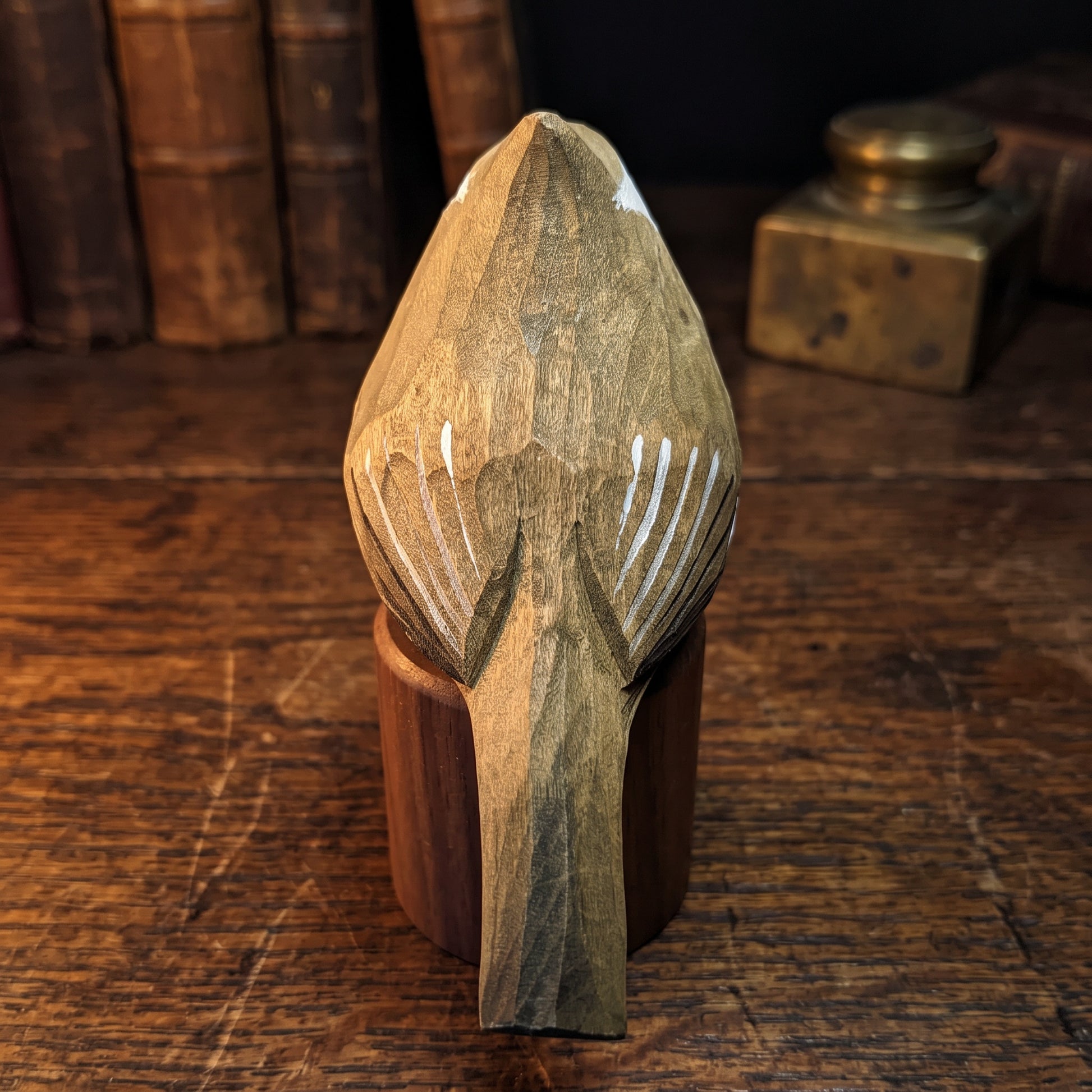 A hand-carved wooden Tufted Titmouse figurine with detailed feather patterns, displayed on a dark wooden stand on a rustic wooden table with vintage books in the background.