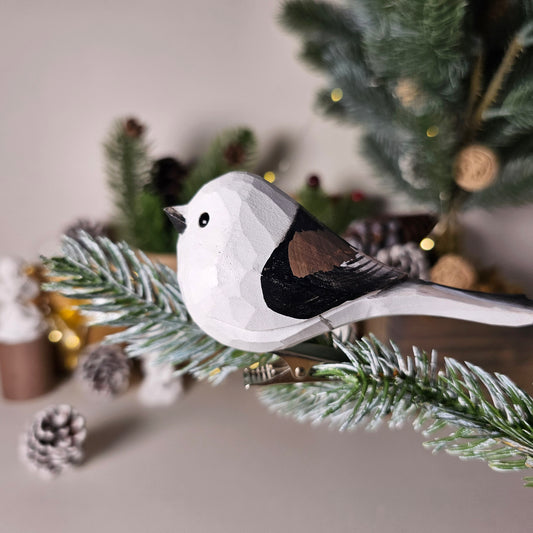 A close-up side-profile photo of a hand-carved wooden Long-Tailed Tit clip-on bird ornament clipped onto a snow-dusted pine branch, featuring a white head with a black eye, a light grey underside, a black and brown back, and a long dark tail, set against a blurred holiday background with pinecones and warm lights.