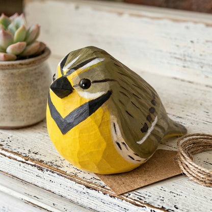 A hand-painted wooden Western meadowlark figurine with a yellow chest, olive-green back, and black beak, resting on a rustic white wooden surface next to a small potted succulent.