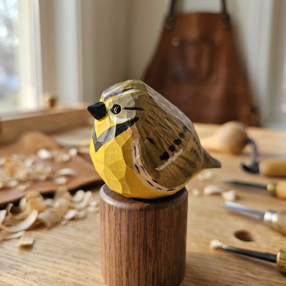 A hand-carved wooden Western meadowlark figurine with yellow, brown, and black painted details, displayed on a small wooden pedestal on a workbench with wood shavings and carving tools in the background.