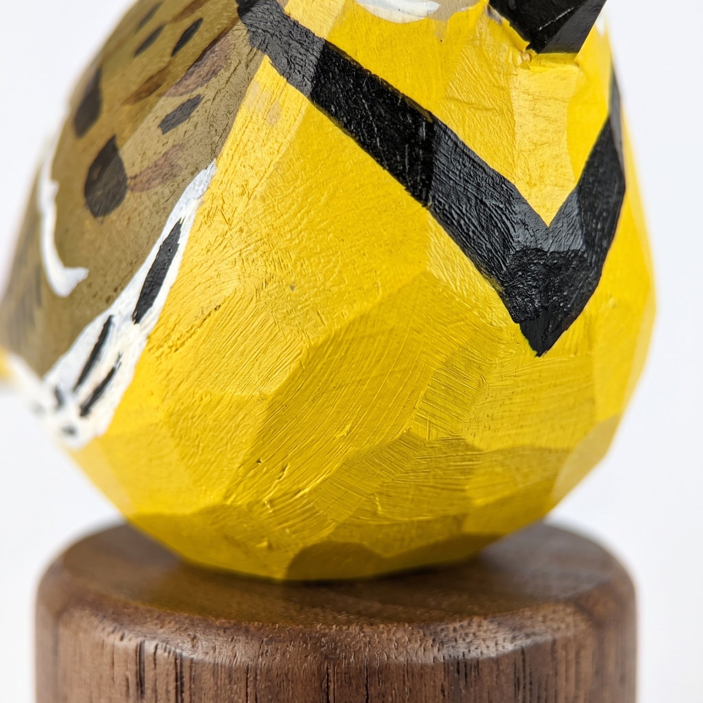 A close-up of a hand-carved wooden Western meadowlark figurine painted bright yellow with black and white markings, mounted on a round wooden base against a white background.