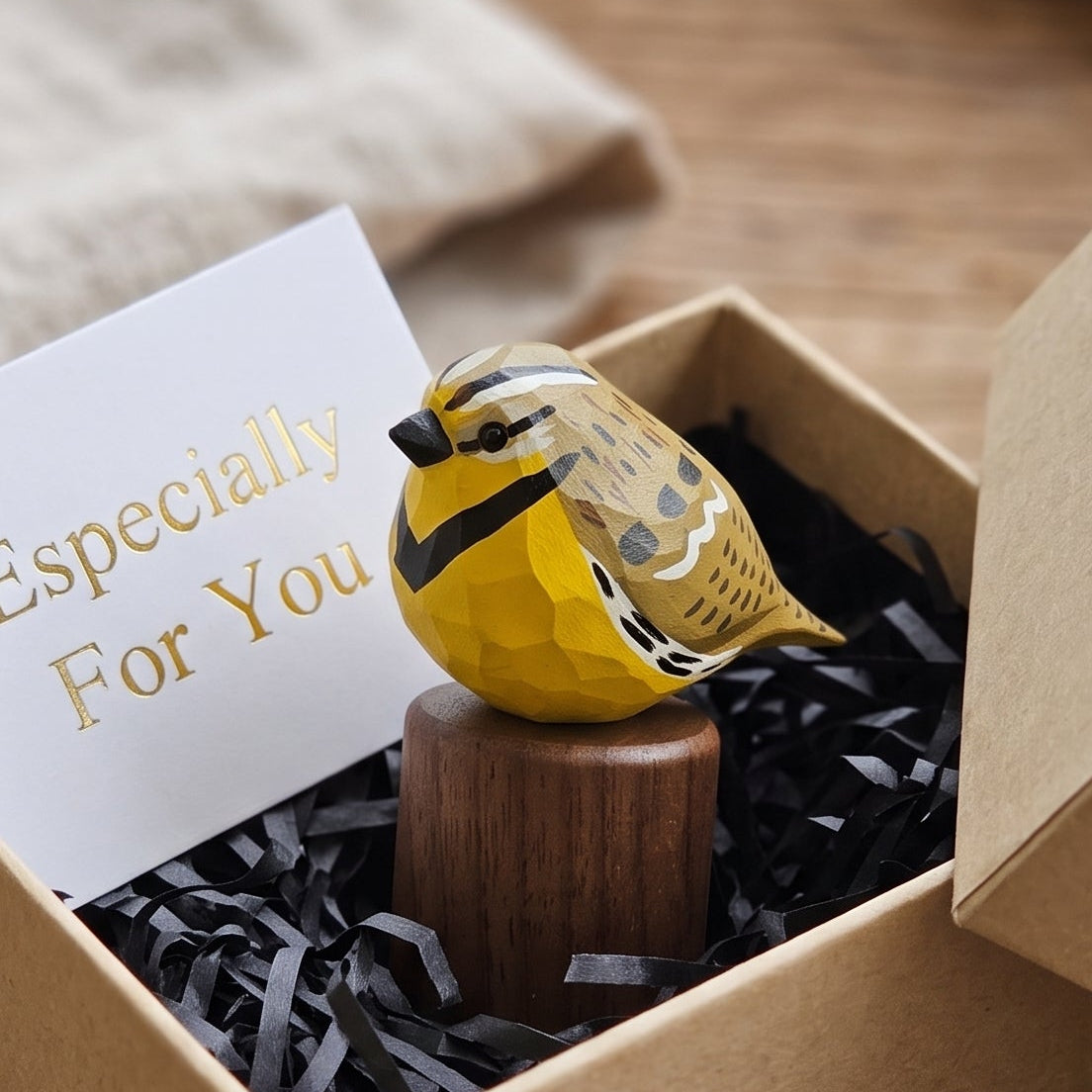 A small yellow Western meadowlark figurine perched on a wooden stand, displayed inside an open kraft gift box with shredded black paper filler and a white card that reads 'Especially For You.' The box sits on a rustic wooden surface with a soft neutral fabric in the background.