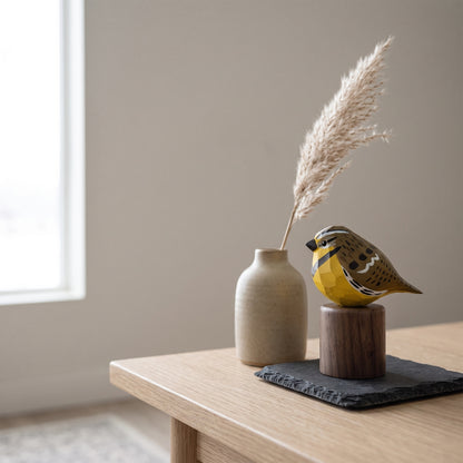 A decorative yellow Western meadowlark figurine with black and brown patterned details sits on a small wooden base next to a beige ceramic vase holding dried pampas grass, arranged on a dark slate tray on a light wood table in a softly lit room.
