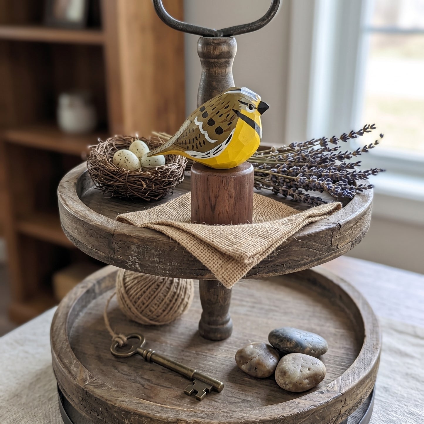 A rustic two-tier wooden serving tray styled with a small yellow Western meadowlark figurine, a nest with eggs, dried lavender, a burlap cloth, a vintage key, and stones, set on a table in a softly lit room.