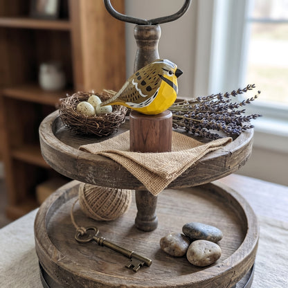 A rustic two-tier wooden serving tray styled with a small yellow Western meadowlark figurine, a nest with eggs, dried lavender, a burlap cloth, a vintage key, and stones, set on a table in a softly lit room.