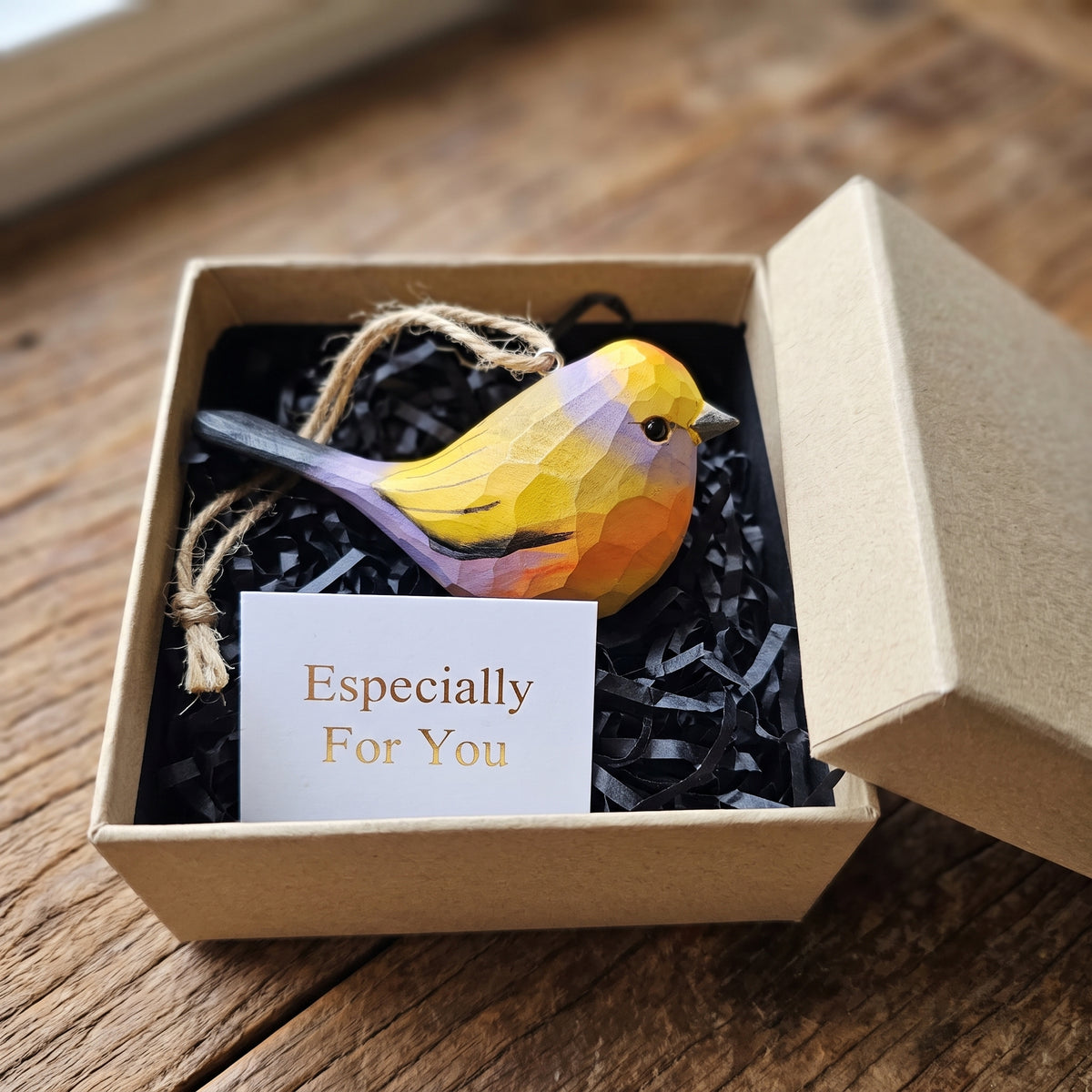 A small White-browed tit-warbler-shaped ornament in yellow, orange, and purple tones, presented in an open kraft gift box with black paper shred and a card that reads 'Especially For You,' set on a wooden surface.