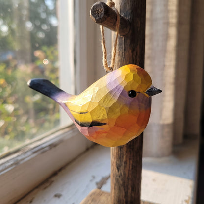 A handcrafted wooden White-browed tit-warbler ornament with a geometric, faceted finish, hanging from a rustic wooden peg on a window sill. The bird is painted in vibrant yellow, orange, purple, and black, set against a soft-focus background of a window and curtains.