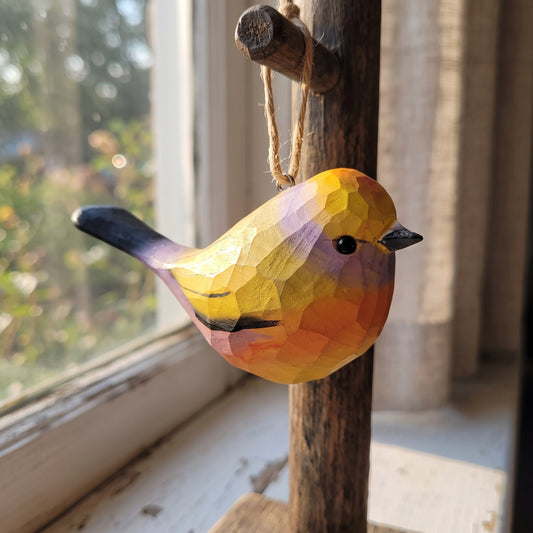 A handcrafted wooden White-browed tit-warbler ornament with a geometric, faceted finish, hanging from a rustic wooden peg on a window sill. The bird is painted in vibrant yellow, orange, purple, and black, set against a soft-focus background of a window and curtains.
