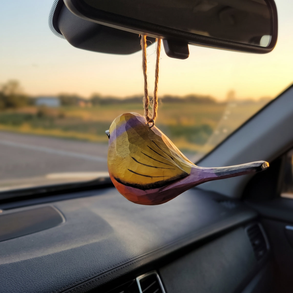 A colorful White-browed tit-warbler-shaped car air freshener ornament hanging from a car's rearview mirror, with a sunset road scene visible through the windshield.