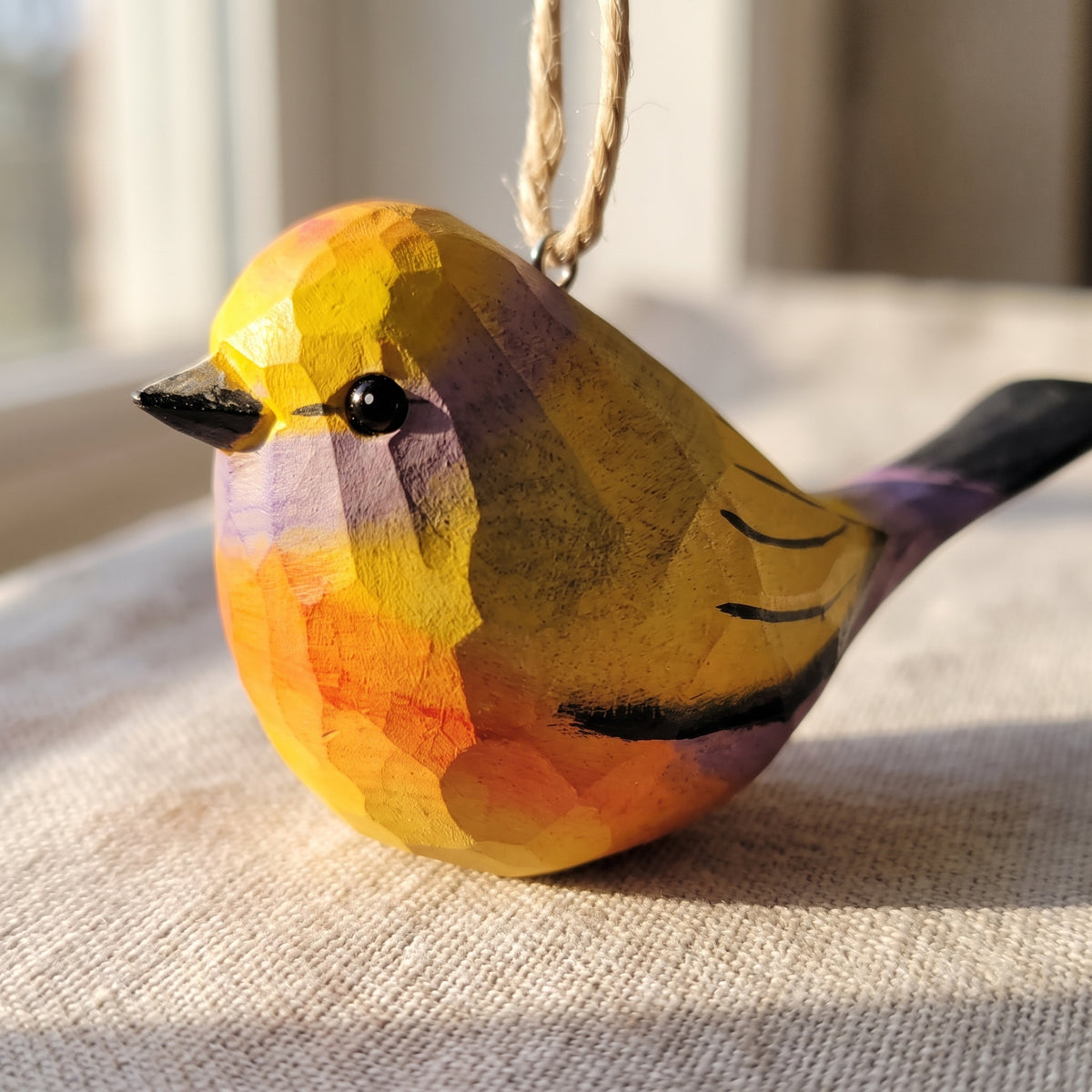 A hand-painted wooden White-browed tit-warbler ornament with a faceted geometric design, hanging by a twine string. The bird features a yellow and orange body with purple and black accents, resting on a textured beige fabric surface.